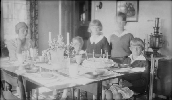 Herbert Edmund Brumder, extreme right, sits at a dining table behind his eighth birthday cake.  His younger brother, Philip, is also seated at the table and his sister Barbara stands beside him.  The child standing between Herbert and Barbara is unidentified.  Their mother, Margaret Bouer Brumder is at the end of the table.  There is a samovar on the side table behind the children.  