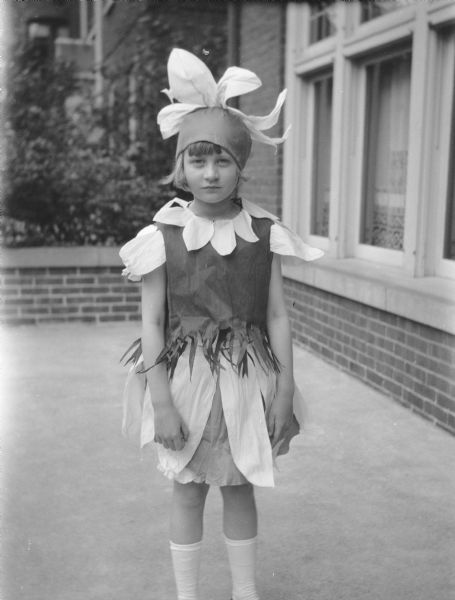 A young girl, probably Barbara Brumder, poses on the porch of a brick house.  She is wearing a costume with a flower shaped hat with large petals.  There are also petals forming her collar and her skirt.