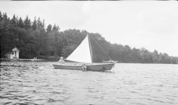 View across water towards Herbert Edmund Brumder. He is using an oar as a tiller on a rowboat fitted with a small sail. A small bathhouse and swimming pier are on the left.