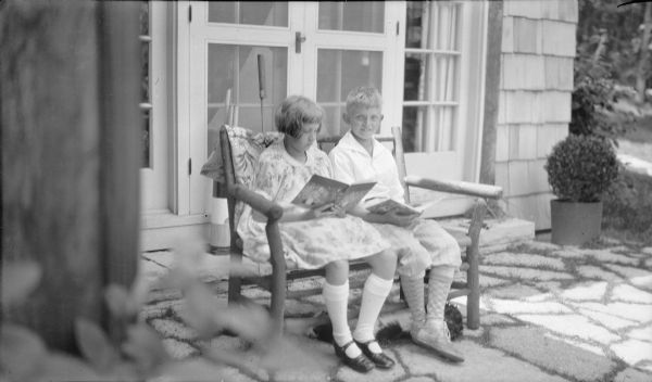 Barbara and Herbert E. Brumder sitting and reading on a rustic bench on a flagstone patio at their summer house on Pine lake. A dog is sleeping under the bench. There are French doors behind them.