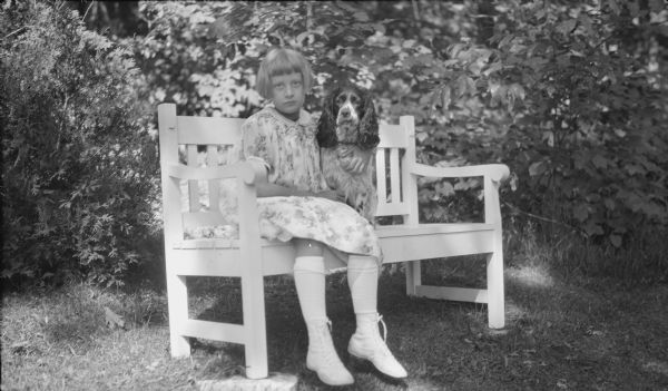 Barbara Brumder, daughter of Herbert P. and Margaret Bouer Brumder, sitting on a garden bench with her arm around a spaniel-type dog.