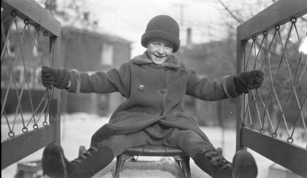 Philip George Brumder, youngest child of Herbert P. and Margaret Bouer Brumder, is smiling broadly while posing sitting on a sled, at the top of a slide in the backyard of the family's home at 2030 East Lafayette Place. He is wearing gloves, a winter coat, stocking cap and galoshes.