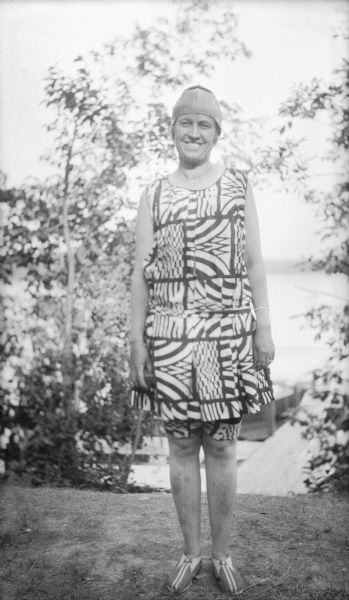 Margaret Bouer (Mrs. Herbert P.) Brumder smiling while posing in a boldly patterned swimsuit. A swimming cap and patterned shoes complete her outfit. A pier and Pine Lake are in the background.