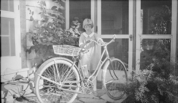 Barbara Brumder, daughter of Herbert P. and Margaret Bouer Brumder, is posing standing with a bicycle on the flagstone patio of the family's summer home on Pine Lake. The bicycle in equipped with a bell on the handle bar and a wire basket mounted behind the seat. The door into the screened porch is behind her.