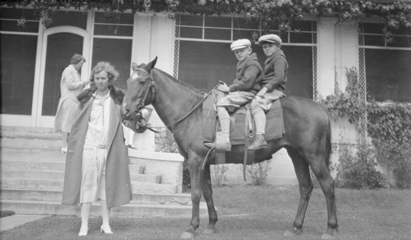 Margaret Bouer (Mrs. Herbert P.) Brumder is holding the reins of a horse ridden by her older son, Herbert E. Brumder (left), and another boy, possibly Herbert's cousin Robert Charles Brumder. Concrete stairs in the background lead to a screen porch. There is an unidentified woman standing on the stairs.