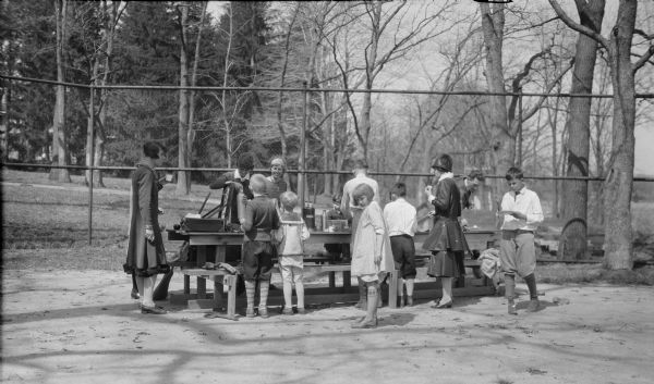 Members of the extended Brumder family are gathered around a long picnic table set up on a tennis court. Built on George and Henriette Brumder's summer estate, Villa Henriette, the tennis court was on a parcel of land inherited by George and Henriette's youngest son Herbert Paul Brumder, who built a house nearby. Herbert's wife, Margaret Bouer Brumder, is seen here on the far side of the table, wearing a necklace and wide headband, smiling at her sons Herbert E., left, and Philip across the table. Daughter Barbara is partially turned to look at the photographer. Pine Lake is on the right.