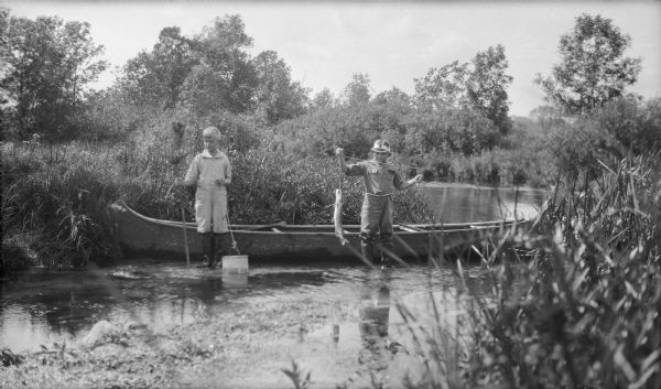 Herbert E. Brumder, left, posing and holding a fishing net in one hand, and the rope to a fishing bucket in the other hand, while standing in front of a moored canoe. An unidentified boy, probably one of Herbert's cousins, is standing on the right holding a fish on a line. The boys are wearing waders and standing in shallow water.