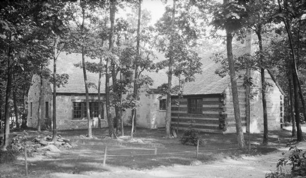 A cottage with a stone wing built at a right angle to a log wing is standing in dappled shade. Between the two wings is a gabled, stone projection. A large stone chimney is centered in the end of the log wing. An area of the lawn is protected by a rope attached to stakes and trees, and marked by a sign that states: "Seeded."