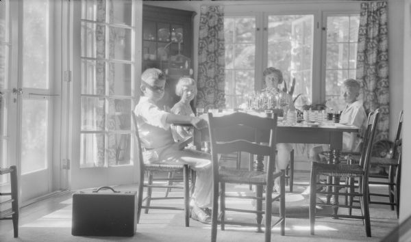 Herbert Edmund Brumder, far left, sitting at a table which is holding a birthday cake with lit candles, and behind it, a floral arrangement in celebration of his tenth birthday. Next to him is his sister Barbara.  Their mother, Margaret Bouer (Mrs. Herbert P.) Brumder is sitting at the end of the table, and at right is Philip George Brumder. Sunlight is streaming in through French doors on the left. There is a china cabinet in the corner and birdcage hanging on a floor stand in front of it. A suitcase is sitting on the floor near Herbert. There is a dog bed under the table.