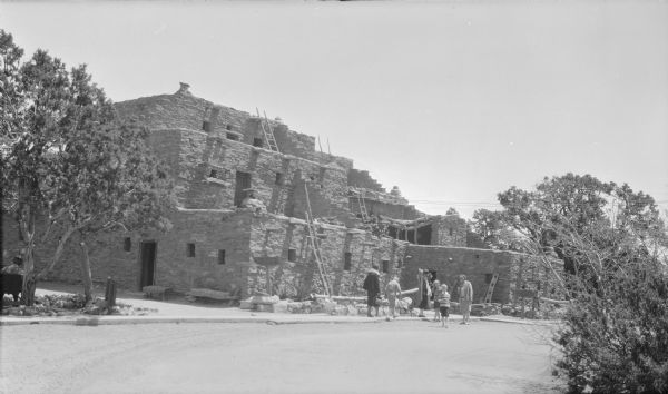 View across road towardsMargaret Bouer (Mrs. Herbert P.) Brumder, far right, posing with two of her children, Barbara and Philip, at a multistory adobe brick pueblo in the southwestern United States.  Multiple ladders lead from one story to another on the three-story structure. There is a small pine tree on the left. A hand-lettered sign at right includes the words: "Purchased in the [illegible] house."