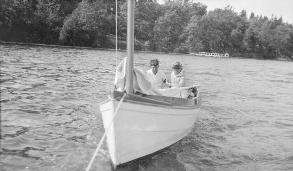 View towards the front of a sailboat in which Herbert E. Brumder, wearing a visor, right, and an unidentified boy on the left, are sitting.  The sailboat which is being towed on Pine Lake. In the background on the tree-lined shoreline is a pier; a birdhouse on a tall pole stands on the sloping lawn.