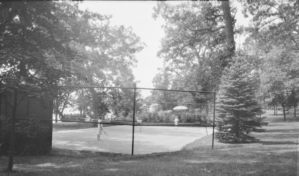 Margaret Bouer (Mrs. Herbert P.) Brumder is standing on a tennis court on the left, opposite her daughter, Barbara, who is wearing a shirt and slacks. There are two shade umbrellas visible beyond the court. At left near the shoreline is a platform with a rustic railing overlooking Pine Lake, with lawn furniture crowded on the platform. A lawn roller is resting at the far end of the tennis court.