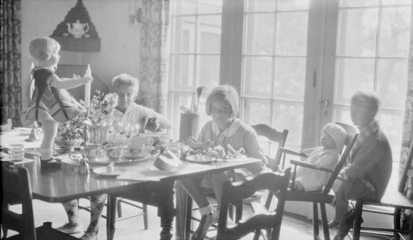 Barbara Brumder, center, is looking at the camera while sitting at a dining table which is set for breakfast. There is a floral arrangement in the center of the table and a cake with lit candles. A large doll with a bold print dress is standing on the table and another doll is sitting in a high chair at Barbara's left. There is a set of golf clubs in a golf bag at her right. Her brothers Herbert E., left, and Philip G., wearing a robe, are sitting nearby. French doors behind the group allow light into the room.