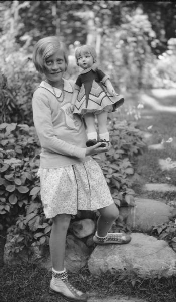 Barbara Brumder, daughter of Herbert P. and Margaret Bouer Brumder, posing standing outdoors holding a large doll, a gift for her tenth birthday. The doll has a short dress with a bold print. Barbara is wearing a sweater and skirt, and is standing with one foot on a stepping stone in the yard.   