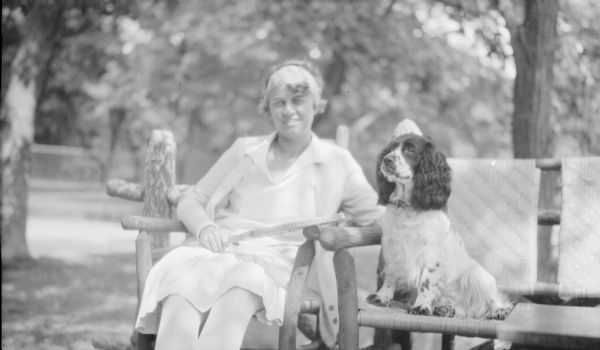 Margaret Bouer (Mrs. Herbert Paul) Brumder posing sitting outdoors on a rustic lawn chair. She is wearing a broad headband, sweater, and short skirt, with long white stockings. She is holding a tennis racquet. A Springer Spaniel is sitting next to her on a rustic settee.