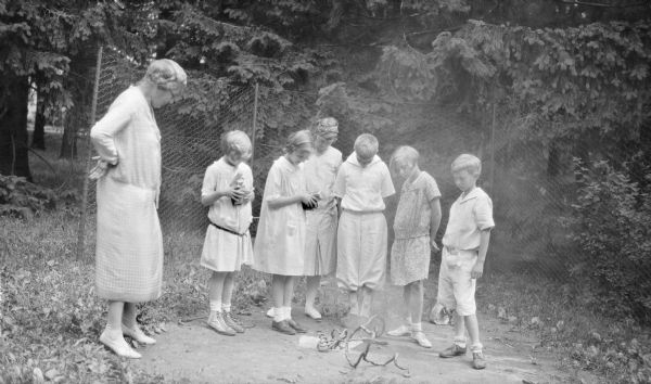 A woman standing on the left, possibly Emma Brumder, and six children are watching a "black snake" type fireworks on a circle of bare ground at Pine Lake. The children, from right, are Philip G. Brumder; Barbara Brumder; and Herbert E. Brumder. The others are unidentified.