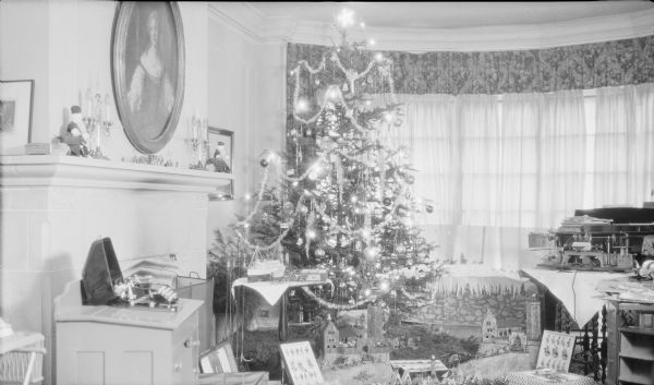 A large Christmas tree, putz (nativity) scene and gifts fill the living room of the Herbert P. Brumder residence, 2030 East Lafayette Place.  The tree is decorated with a variety of ornaments, tinsel and electric lights. There is a small typewriter in an open case at left. On the right is a small lathe, drill press and saw. On the floor in front of the tree, small toy soldiers defend two miniature walled cities with castles and towers.