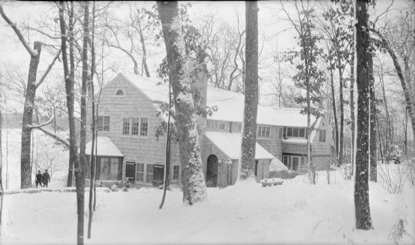 An elevated winter view looking downshill towards the shingle style summer home of Herbert P. Brumder, the youngest son of George and Henriette Brandhorst Brumder. There is a large bay window on the left side of the house, and a stone extension at right includes the arched front door and a large chimney. Two children are standing at the far left side of the house, with Pine Lake in the background. The house was built on land that was part of the original Brumder country estate, Villa Henrietta.