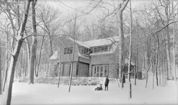 A winter view of the lake-facing side of Herbert P. Brumder's shingle style summer house on Pine Lake. Two children, one lying on a sled, are posing in the foreground. The screened windows of the porch have been covered for the winter. The house was built in 1926 on land which was originally part of George and Henriette Brumder's summer estate, Villa Henrietta.