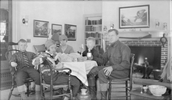 The Herbert P. Brumder family enjoying a wintertime lunch in their Pine Lake house. They are, from left, Herbert E., Barbara, Margaret, Philip, and Herbert P. Brumder. Barbara and her mother are wearing sweaters; Herbert E. and his father are wearing woolen shirts and knickerbockers with high stockings. There is a fire in the fireplace. An Audubon print featuring canvasback ducks is hanging over the mantle. A large Thermos type container is sitting on the floor, and Herbert P. is holding a tin cup.
