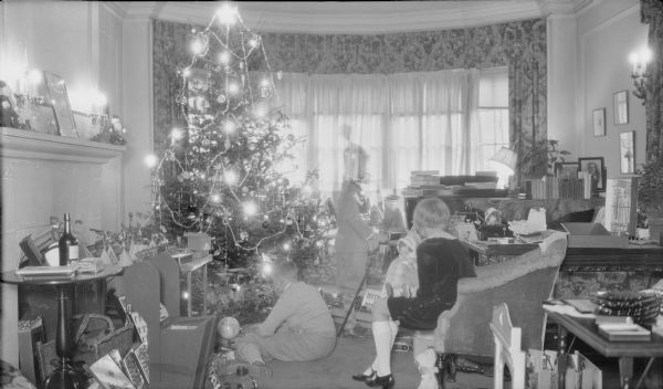 The three children of Herbert P. and Margaret Bouer Brumder, from left, Herbert E., Philip (partially obscured), and Barbara are playing with their Christmas presents in the living room of their home at 2030 East Lafayette Place. Barbara, sitting in an armchair, is holding a doll. On a table at left is a miniature military encampment with detailed metal cannons; a small world globe is resting on the floor. The electrically lighted Christmas tree is decorated with tinsel garland and glass ornaments. At far right, a photograph of Milwaukee publisher and businessman George Brumder is sitting on the grand piano. Herbert P. was the youngest child of George and Henriette Brandhorst Brumder.