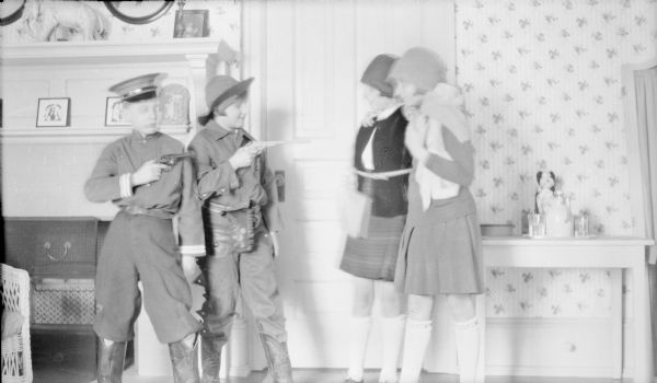 Two girls dressed as flappers with cloche hats, right, are being held at gunpoint by a boy and girl dressed as outlaws wearing cowboy boots. The boy is Philip Brumder; the girls are not identified. There is a fireplace with tile surround at left, and a small table with pitcher and glasses on the right.