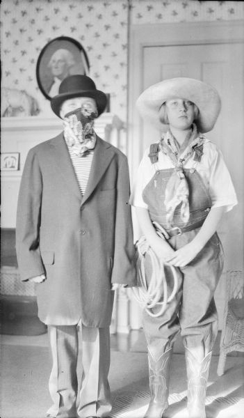 Herbert E. Brumder is dressed in an oversized overcoat, slacks and hat, and is wearing a bandana face mask. He is standing next to his sister Barbara, who is dressed as a cowgirl. She is wearing a large-brimmed hat, neckerchief, bib overalls and western boots. She has a rope coiled at her waist.