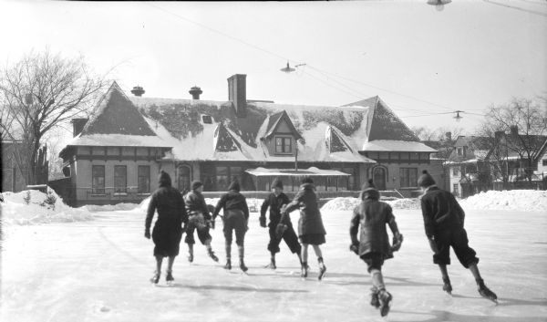 Seven children are ice skating toward the rear of a long, one-story building with half timber details under the eaves and a massive central chimney. Piles of snow line the large rink. Snow has been cleared up to the building, which appears to be a commercial building, or possibly a clubhouse. Electric lights are hanging from wires suspended over the rink. There are houses along a street in the background on the right. 