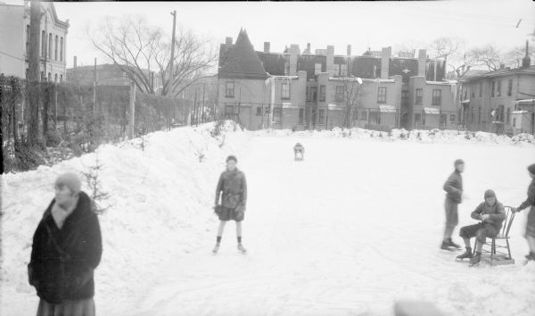 The photographer's wife, Margaret Bouer Brumder, is standing in the foreground on the left, as her daughter, further back, is skating toward her. There are other children on the ice, including a boy sitting on a chair at right. Small evergreen trees are protruding from the snow piles around the perimeter of the rink. In the background is the rear facade of a block of three-story row houses which have a two-story rear extension. Beyond a fence at left is a tall brick commercial building; there are other buildings in the background.