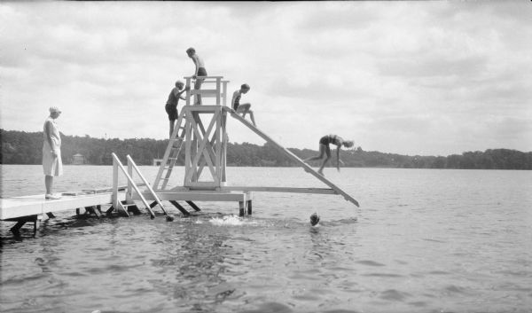 View across water towards Margaret Bouer (Mrs. Herbert P.) Brumder, wearing a light-colored dress and cap, standing on a pier observing as children are playing on a water slide on Pine Lake. There are two children in the water swimming toward the ladder. A diving board extends from the end of the pier. An octagonal boathouse is on the far shoreline.
