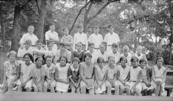 Group portrait of boys and girls posing on a long picnic table near the summer home of Herbert P. and Margaret Bouer Brumder on Pine Lake. The children are wearing light-colored summer clothes, and some of them have hair damp from swimming. The photographer's children can be identified: Herbert E. Brumder is in the middle row, far left; his brother Philip is also in the middle row, second from right. Their sister, Barbara is in the front row, far left. The group also includes cousins who are the grandchildren and great-grandchildren of Milwaukee publisher and businessman George Brumder and his wife Henriette. 