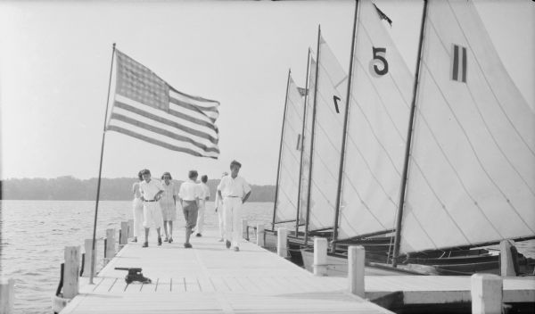 Five dinghy sailboats are moored to the right side of a wide pier on Pine Lake. Several unidentified young men and women are walking on the pier. There is a yacht cannon on the pier in the foreground, near a flag pole on the left flying a 48 star flag. There is a shoreline in the distance.