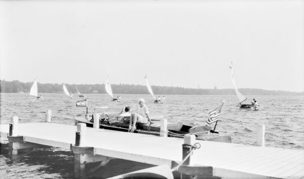 A young woman, possibly the photographer's daughter Barbara Brumder, is posing sitting on the deck of a wooden Chris-Craft power boat moored to a pier. There are flags fore and aft on the power boat. An unidentified man is sitting in the boat, watching a dinghy sailboat race. Each of the boats has a two-person crew.  A flag flies from the top of an ornate observation tower visible on the far (eastern) shore of Pine Lake.  