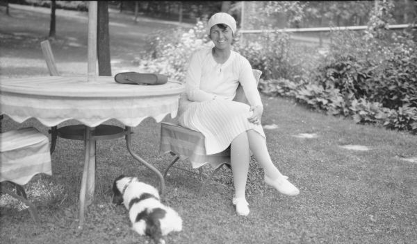 The photographer's wife is  posing sitting at a round outdoor table near their summer home on Pine Lake. She is wearing a light-colored dress and cap. The table and chairs have striped canvas covers and there is a camera case on the table. In the background is a path of stepping stones along a flower border and tall fence. The family's Springer Spaniel is resting on the grass under the table.