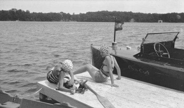 Barbara Brumder, right, lounging on a pier and watching as an unidentified girl next to her is petting the Brumder family Springer Spaniel. The girls are wearing swimming suits and caps. An oar and a yacht cannon is resting on the pier, and the side of a rowboat is in the left foreground. There is a Chris-Craft power boat moored to far side of the pier.