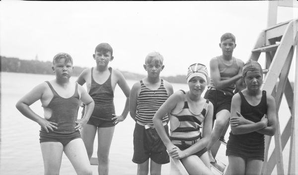 Six children, four boys and two girls are posing near the diving platform on a swimming pier on Pine Lake. The children, who are wearing bathing suits, are likely all members of the extended Brumder family. The girl wearing the striped top and cap is Barbara Brumder; her younger brother Philip, behind her, is wearing a top with narrow stripes. Barbara and Philip are children of Herbert P. and Margaret Bouer Brumder.