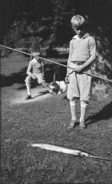 Herbert E. Brumder posing standing and holding a fishing pole and line. A longnose, or needlenose gar is lying on the ground in front of him. Herbert's younger brother Philip is sitting on a swing in the background, with the family dog near him.