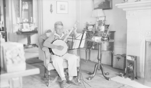 Philip George Brumder posing with a banjo, sitting in an easy chair in the living room of the family home at 2030 East Lafayette Place. There is sheet music on the music stand in front of him. The fireplace at right has a carved stone surround.
