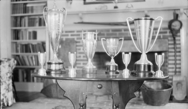 Eight loving cup trophies of various sizes standing on a gate leg table in the living room of Herbert P. Brumder's summer home on Pine Lake. Although the inscriptions on the cups are not entirely legible, several include an inscription of the Pine Lake Yacht Club. The tall cup second from left is identifiable by its shape as the Robert E. Friend Trophy which was won by the photographer's son Herbert E. Brumder in 1929.