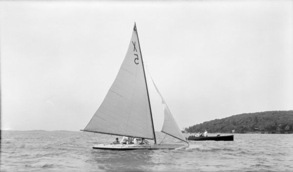 View across water towards a crew working on a racing yacht identified as X5 as a team of judges in a wooden powerboat observes. A tree-lined shoreline is on the right.