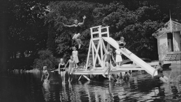 View across water towards a group of boys and girls, likely all members of the extended Brumder family, playing and swimming near a water slide at the end of the swimming pier at Villa Henrietta, the summer estate on Pine Lake established by Milwaukee publisher and businessman George Brumder. Herbert E. Brumder appears to hover over several other children while jumping into the water. A boathouse with intricate gable fretwork and a small spire is on the shoreline on the far right. There is a canoe on the pier.