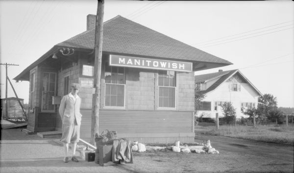 Herbert E. Brumder standing near a suitcase, large bag and other parcels resting on the ground at the small Chicago Northwestern Railroad depot at Manitowish. There is a house across a lane behind the depot. A large sign identifies the location, and there is small sign advertising Western Union services.