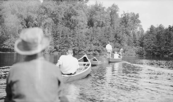 A man standing in a small boat controls a trolling motor with a rope as he leads a flotilla of canoes. There is a densely wooded shoreline in the background. There are two women in a canoe next to the lead boat. A young man, probably Herbert E. Brumder, is paddling in the third canoe. The photographer is behind a man sitting in the boat in the left foreground.