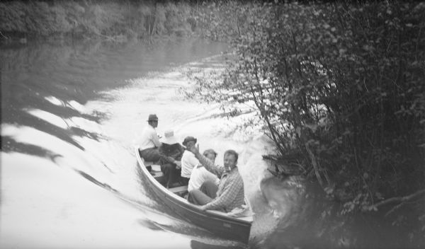 Elevated view of a man smoking a cigarette sitting in the bow of a motor propelled wooden boat waving a greeting toward the photographer. There are four other persons in the boat, which is making a turn around a wooded point, possibly into an inlet.