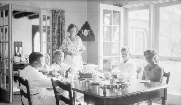 Birthday boy Herbert Edmund Brumder, second from right, is sitting at a dining table with his siblings Barbara, right, and Philip, who is seated at the head of the table. Another young man, probably a cousin, sits at left. Herbert's mother, Margaret Bouer Brumder, is standing behind the table, and is wearing a housecoat with ruffles at the lapesls and sleeves. There is a birthday cake and a box of new golf balls on the table in front of Herbert. Morning sunlight is streaming through the open French doors from behind the table on the right. A room with a beamed ceiling is through a doorway on the left. The celebration is at the Brumder's summer home at Pine Lake.