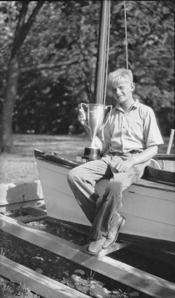 Herbert E. Brumder, son of Herbert P. and Margaret Bouer Brumder, is sitting on the deck of his sailboat holding the Pine Lake Yacht Club championship trophy for his class. The boat is secured on rails near the Brumder family's summer home on Pine Lake.