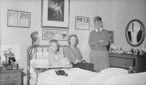 A metal overbed table holding a birthday cake and wrapped gift has been positioned over the bed of Philip Brumder, who sitting up in bed and is wearing a robe. His sister Barbara is sitting on the bed beside him, and brother Herbert E. is standing at right. There are 10 lighted candles on the cake. Framed prints are hanging  on the wall behind the bed.  