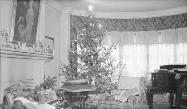 An electrically lighted Christmas tree is standing near the bow window in the home of Herbert P. and Margaret Bouer Brumder at 2030 East Lafayette Place. The tree is trimmed with tinsel and glass ornaments.  Wrapped gifts are piled in a chair near the tree. There is a toy power boat and erector set at left, near a poinsettia plant. A portrait of the Brumder's daughter Barbara with the family Springer Spaniel is hanging over the fireplace. At right are a grand piano and radio.