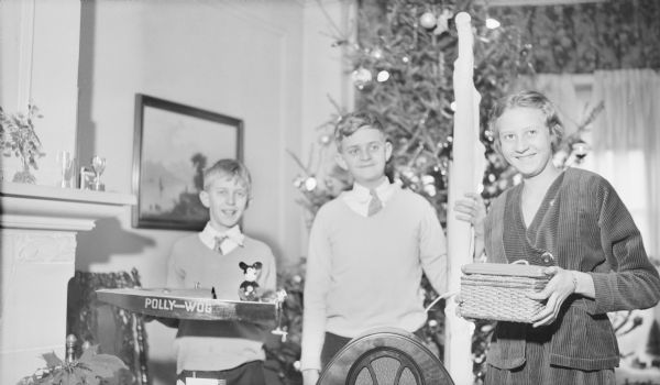The three children of Herbert P. and Margaret Bouer Brumder are smiling as they are posing standing in front of the Christmas tree holding gifts. From left, Philipis holding a toy power boat with a Mickey Mouse figure in it; Herbert E. is standing and holding what appears to be a furled sail; and Barbara is holding a basket with a long narrow box resting on it. There is a poinsettia plant at lower left. The top of a radio is in the foreground.