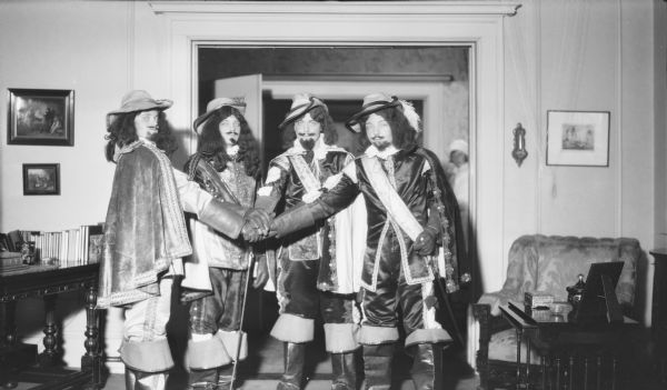 Four young men are dressed in elaborate costumes as the Three Muskateers and D'Artagnan and are posing in the Herbert P. Brumder house at 2030 East Lafayette Place. Each man has placed his right hand over the hilt of a sword in the center of the group. A woman wearing a hat and coat is visible through the doorway in the background. Written on the negative envelope is: "Dress-up Skating Party."